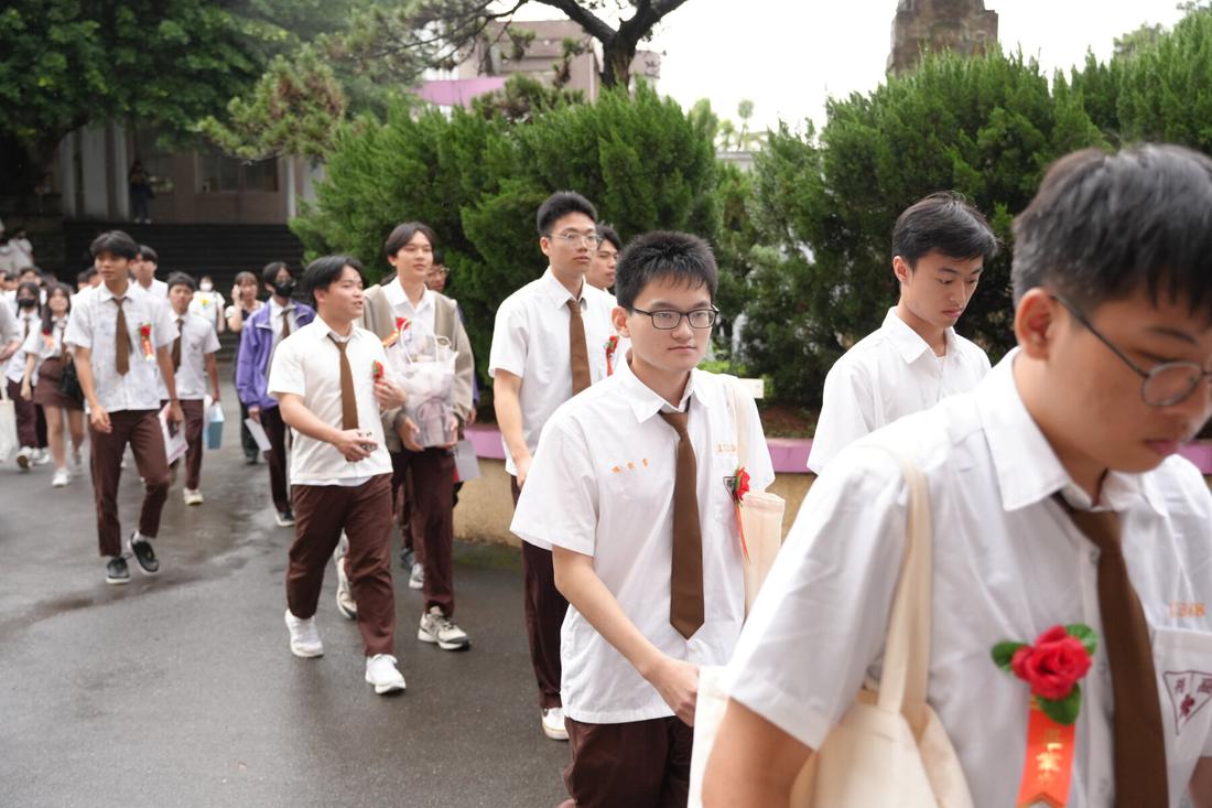 畢業生沿校園步道行進，衣領別著紅色胸花。